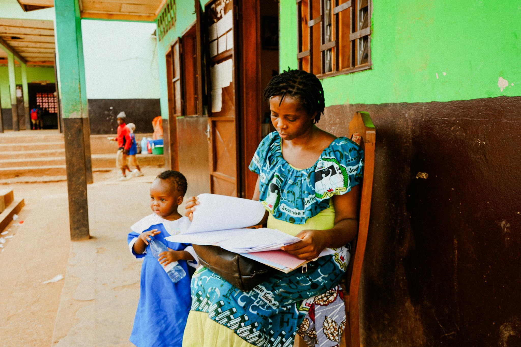 A mother reading documents while her child stands beside her in a school setting.